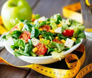 Fitness salad and measuring tape on rustic wooden table. Mixed greens, tomatos, diet cheese, olive oil and spices for healthy lifestyle concept.